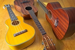 Collection of three ukuleles aligned on sun drenched wooden table top. High angle view. Side lit, showing texture of wood grains.