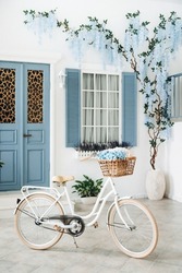 Vintage white bike with flowers in a basket against the blured background of a white mediterranean house with a blue door and window and flowering tree on the wall.