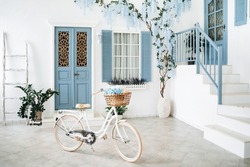 Mediterranean style white house exterior with blue door and window, blossoming tree and white vintage bike with flowers in a basket and. Traditional patio of Santorini