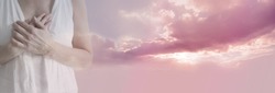 True Compassion from my Heart - female in white tunic with hands crossed over heart fading into a beautiful gentle pink clouded sun set with copy space on right side
