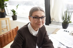 Close up portrait of happy beautiful 60 year old experienced female chief architect wearing jacket and eyeglasses doing paperwork in office, smiling joyfully at camera, enjoying her occupation