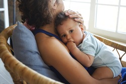Indoor picture of unrecognizable young dark skinned female sitting in armchair, holding cute adorable toddler, stroking his hair. Lovely infant resting in mother's hands, looking at camera
