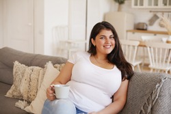 Beautiful plus size woman in casual clothes sitting on couch in living room with cup of morning coffee, smiling, thinking about new day, making plans. People, lifestyle and domesticity concept