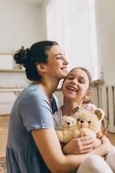 Smiling mother hugging cute little girl holding teddy bear toy, both laughing while cuddling, girl sitting on mom's knees, female tickling her child. Human relationships. Carefree childhood