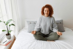 Pretty woman practicing yoga at home. Portrait of beautiful redhead caucasian plus-size female with curly hair sitting on bed in lotus posture, relaxing her mind, doing spiritual practice