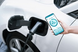 Battery status of electric vehicle displayed on smartphone application or software while vehicle is plugged into charging station for progressive future refueling. Battery status on phone screen.