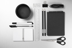 Black and white stationery on gray desk: notepads, pencils, scissors, stapler, clip, pins, ruler, pens, pen pot. Top view point.