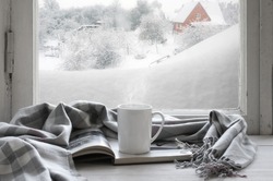 Cozy winter still life: mug of hot coffee and opened book with warm plaid on vintage windowsill of cottage against snow landscape with snowdrift from outside.