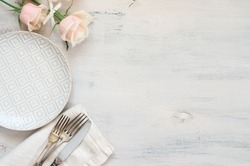 Light pastel colored tableware set: plate, vintage silverware on napkin and delicate pink roses on rustic shabby wooden background with copy space. Top view.