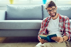 Portrait of joyful young man reading book while sitting on floor in his living room