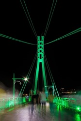 Perspective view of illuminated span and pylon of the Bridge of Lovers with people blurred in motion at night, Tyumen, Russia
