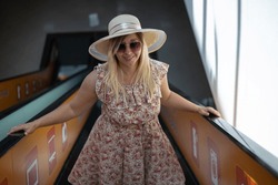 A cute smiling woman lifts up the escalator in a sundress in a shopping mall. On a hot summer day, a woman went shopping in a cool mall. Concept woman summer in the mall.