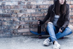 Young woman in hood is sitting on skateboard and holding smoldering cigarette and bottle of beer. Homeless teenage girl is drinking and smoking in abandoned building. Dregs of society concept.