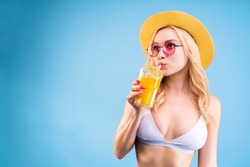 Young woman in swimsuit is having rest, relax, leisure pause from work concept. Photo studio shooting portrait of attractive lady is tasting yummy cocktail from glass isolated on blue background. 