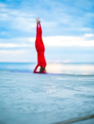 blured, female in a red yoga suit makes a headstand on a yoga mat near a sea, blue clouds on the horizon