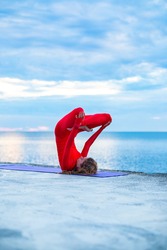female in a red yoga suit makes a headstand asana on a yoga mat near a sea, blue clouds on the horizon