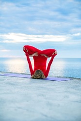 female in a red yoga suit makes a headstand asana on a yoga mat near a sea, blue clouds on the horizon