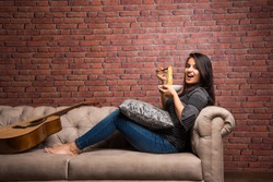 Young beautiful asian/indian girl eating instant noodle using chopsticks while seating at sofa at home, selective focus
