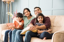 Portrait of happy Indian Asian young family while sitting on sofa, lying on floor or sitting against wall