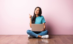 Indian asian young woman or girl sitting with laptop on her lap against pink wall on wooden floor