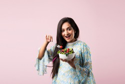 Portrait of a happy Indian asian pretty young woman eating fresh salad from a bowl standing isolated over yellow background
