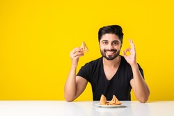 Handsome Indian bearded young man eating Samosa snacks while sitting at white table against yellow background