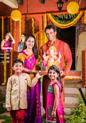 Happy Indian Family Celebrating Ganesh Festival or Chaturthi - Welcoming or performing Pooja and eating sweets in traditional wear at home decorated with Marigold Flowers