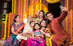 Happy Indian Family Celebrating Ganesh Festival or Chaturthi - Welcoming or performing Pooja and eating sweets in traditional wear or taking selfie picture at home decorated with Marigold Flowers