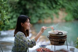 Thai style barbecue ,Young asian woman enjoy eating and grilling pork or meat on hot pan with nature waterfall view background. Moo-Kata in Thailand.