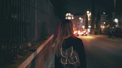 Back view of brunette woman walking near the road at the traffic time. Girl goes through the city late at night alone.