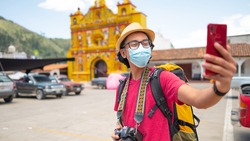 Portrait of a tourist with a medical mask, backpack, and camera taking a selfie with a cell phone.