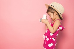 Portrait of cute little girl in straw hat and pink jumpers taking picture with white camera in the studio on pink background. Photography concept. Copy space for text