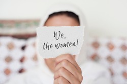 closeup of a young woman indoors wearing a white hijab showing a piece of paper in front of her face with the text we the women written in it