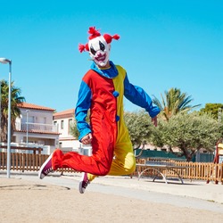 a scary clown wearing a colorful yellow, red and blue costume, jumping in an outdoor public playground