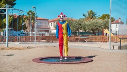 a scary clown wearing a colorful yellow, red and blue costume, bouncing on a trampoline in an outdoor public playground