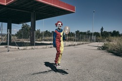 a scary clown wearing a yellow, red and blue costume, holding a golden balloon in his hand and jumping in front of an abandoned filling station
