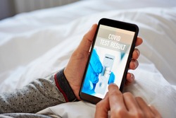 closeup of a young caucasian man in pajamas, lying in bed, checking the result of his covid test in a simulated app in his smartphone, that reads covid test result