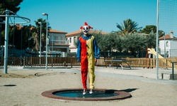 a creepy clown, wearing a colorful yellow, red and blue costume, is bouncing on a trampoline in an outdoor public playground