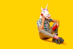 a young man wearing a unicorn mask talking on the phone, using a colorful red landline telephone, sitting on a yellow background