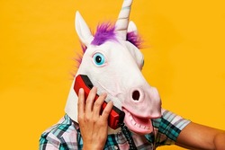 closeup of a young man wearing a unicorn mask talking on a colorful red landline telephone on a yellow background