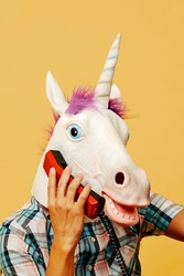 closeup of a man wearing a unicorn mask talking on a colorful red landline telephone on an orangish yellow background
