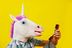 closeup of the head of a young man wearing a unicorn mask, talking on a red landline telephone, against a yellow background