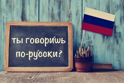 a chalkboard with the question do you speak russian? written in russian, a pot with pencils, some books and the flag of Russia, on a wooden desk