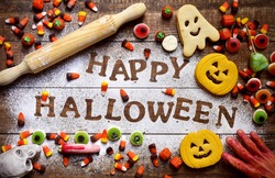 high-angle shot of a wooden table sprinkled with icing sugar where you can read the text Happy Halloween, some different candies and cookies, some scary ornaments and a rolling pin