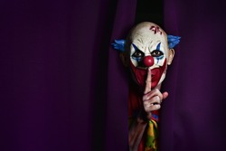 a scary evil clown peering out from a purple stage curtain, with his forefinger in front of his lips, asking for silence, with a negative space on one side