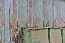 Paint chipped, weathered wood wall with wooden boxes in front of it. Aqua-green and aqua-blue paint chipping of and wood damp and rotting. Picture taken at an angle to the wall.