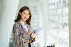 Business woman in light jacket stands and writes something. Asian girl writes with pen in note goals plans affairs and list of wishes for work. Notes of successful lady.