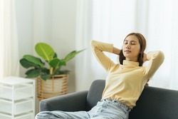 Young attractive beautiful asia female girl or university student sit smile look outside window put arm hand back behind head at sofa couch living room feeling relaxed comfort at cozy home.
