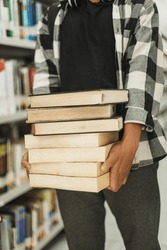 Close-up hands carrying big stack of books against the background of bookshelves in a library. Learning concept, lifestyle. 