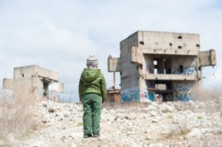 one little child in green jacket standing on ruins of destroyed buildings in war zone